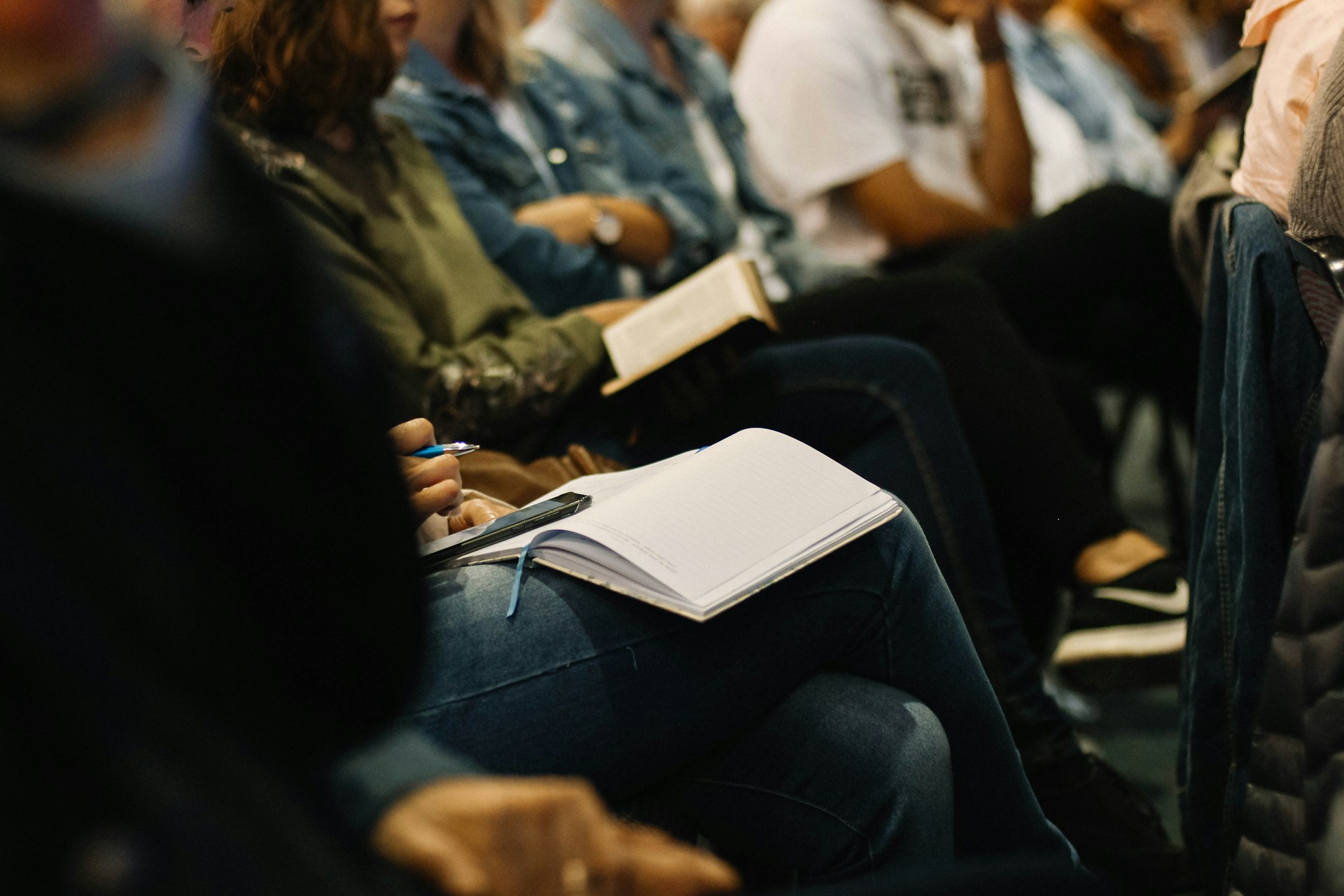 Une rangée de personnes participant à une discussion de type assemblée publique, écoutant un conférencier avec des cahiers sur les genoux. | A row of people at a Town Hall-style discussion, listening to a speaker with notebooks on their laps.